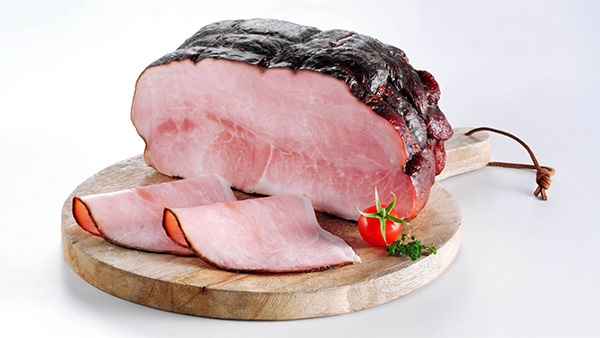 Serviervorschlag: Kernrauchschinken auf Holzbrett mit zwei aufgeschnittenen Scheiben, dekoriert mit Tomate und Petersilie.
