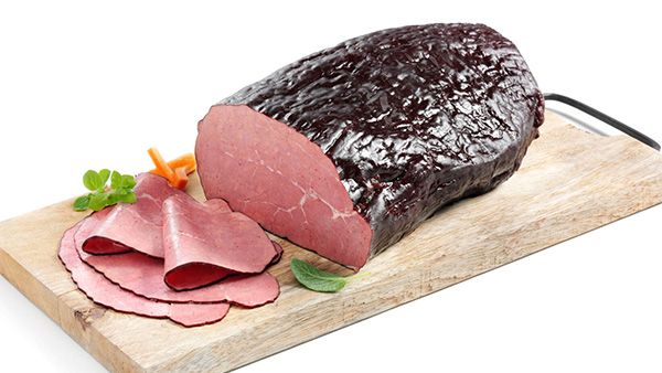 Serviervorschlag: Rindersaftschinken auf Holzbrett mit zwei aufgeschnittenen Scheiben, dekoriert mit Tomate und Petersilie.